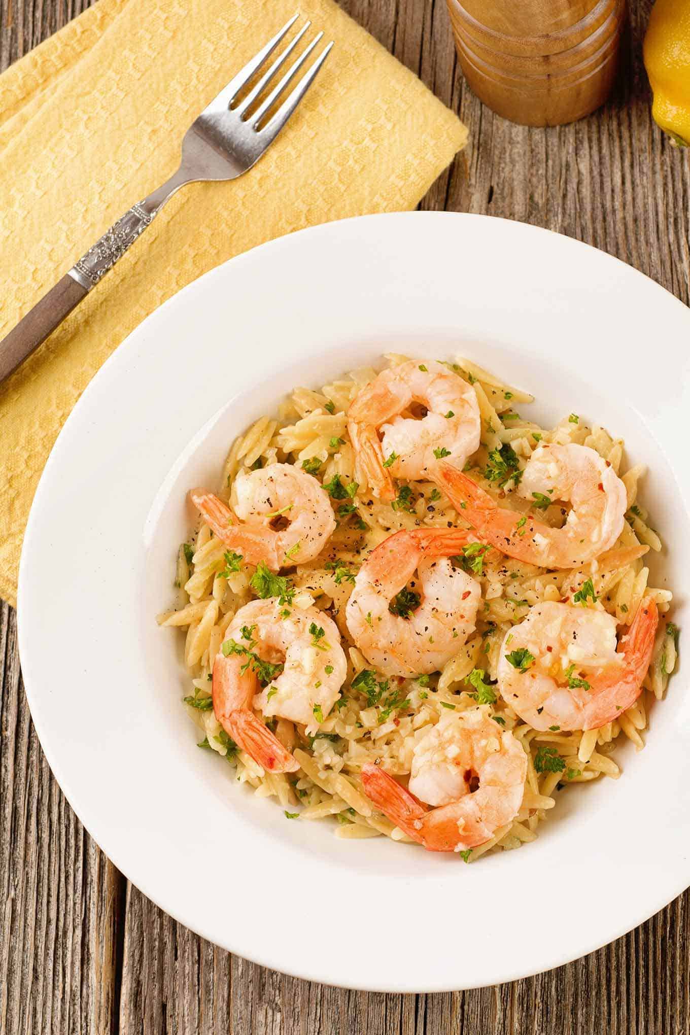 A bowl of sautéed garlic butter shrimp served over orzo cooked in chicken broth, with parsley garnish, a fork and napkin on the side.