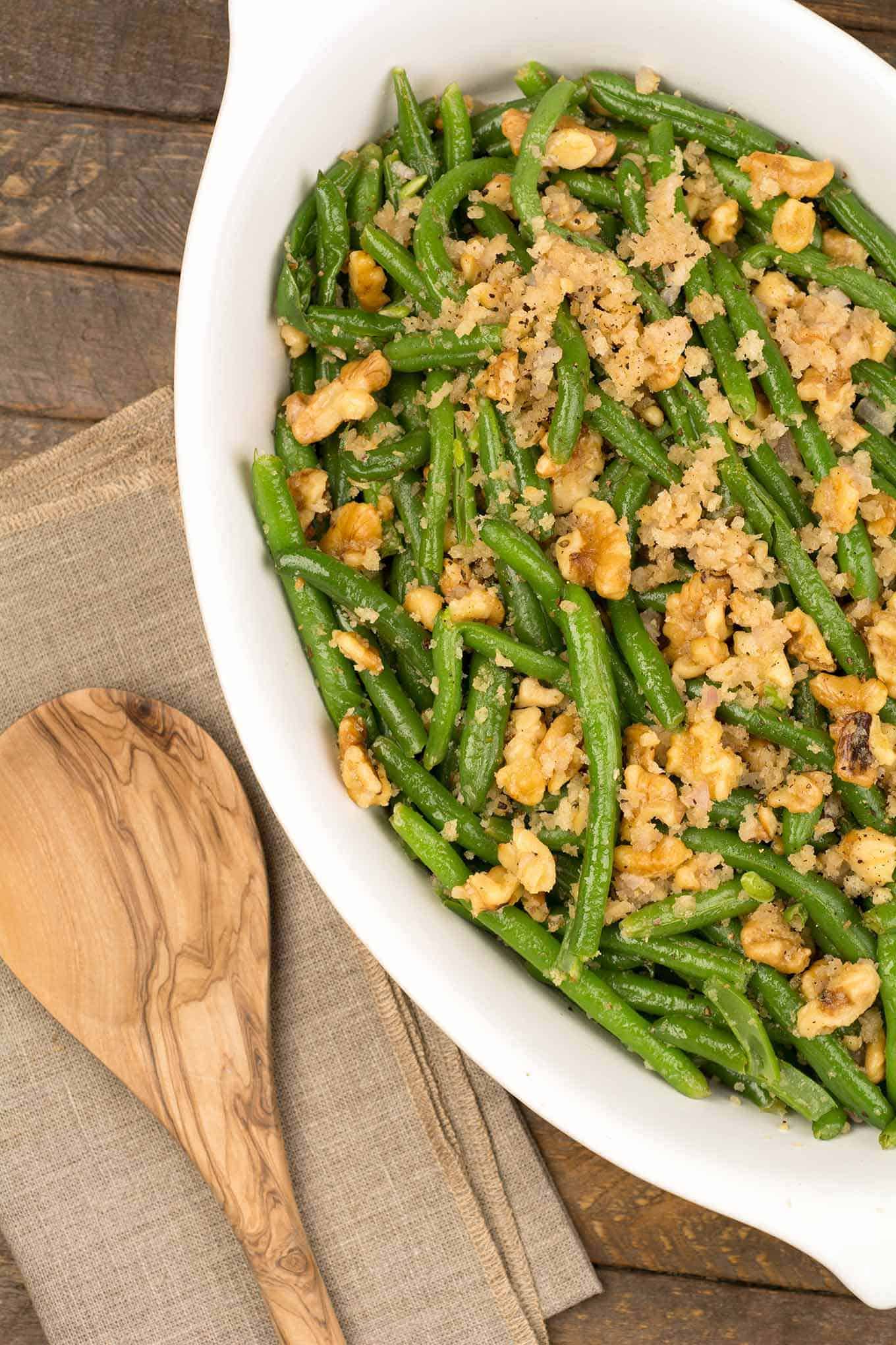 A white oval casserole of roasted fresh green beans with a crispy topping of toasted walnuts and sautéed shallots, a wooden spoon to the left.