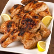 Grilled spatchcocked, honey-glazed Cornish hens with charred skin on a white platter with lemon wedges on the side.