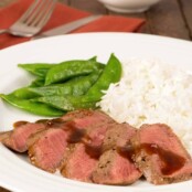 A plate of sliced, grilled flat iron steak drizzled with orange-hoisin sauce and served with steamed rice and snow peas, napkin and utensils in the background.