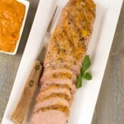 An oblong platter with a partially carved, grilled pork tenderloin topped with peach barbecue sauce, serving fork and extra sauce on the side.