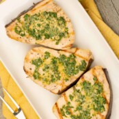 A serving plate with three portions of grilled swordfish steaks topped with lemon basil butter, a grilling fork to the left and napkin beneath the plate.