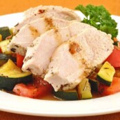 Sliced herb and butter poached chicken breast served over a medley of sautéed zucchini and red bell peppers, garnished with parsley on a white plate.
