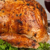 Mayonnaise and herb coated turkey with golden brown skin, ready for carving, on a platter garnished with fresh herbs, a glass of red wine in the background.