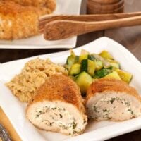 Rolled, crumb-coated chicken breasts filled with herbed goat cheese, served with sautéed zucchini and orzo on a square white plate, more chicken in the background.