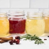 Four glass mason jars filled with homemade holiday simple syrups orange, cranberry, rosemary, and ginger, ingredients scattered in front.