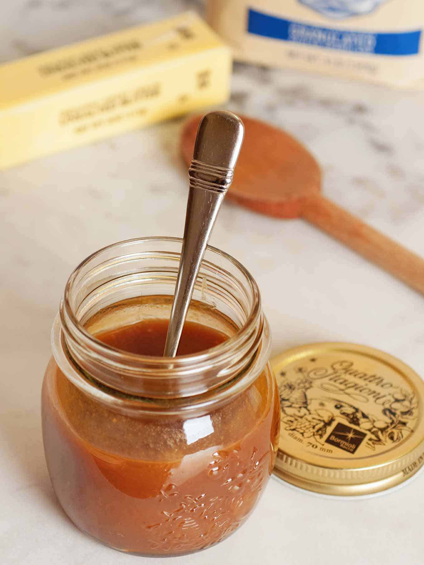 Homemade caramel sauce in a clear jar with spoon, lid, wooden spoon, stick of butter and sugar in the background.
