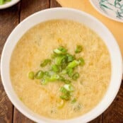 An overhead view of a bowl of egg drop soup garnished with scallions, a dish with more scallions, a napkin, and a ceramic spoon in the background.