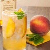 A peach and honey whiskey cocktail in a collins glass, garnished with peach slices and fresh mint. A whole peach and a bottle of whiskey in the background.
