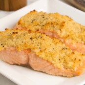 Two fillets of roasted salmon with a golden horseradish and crumb crust on a white square plate.