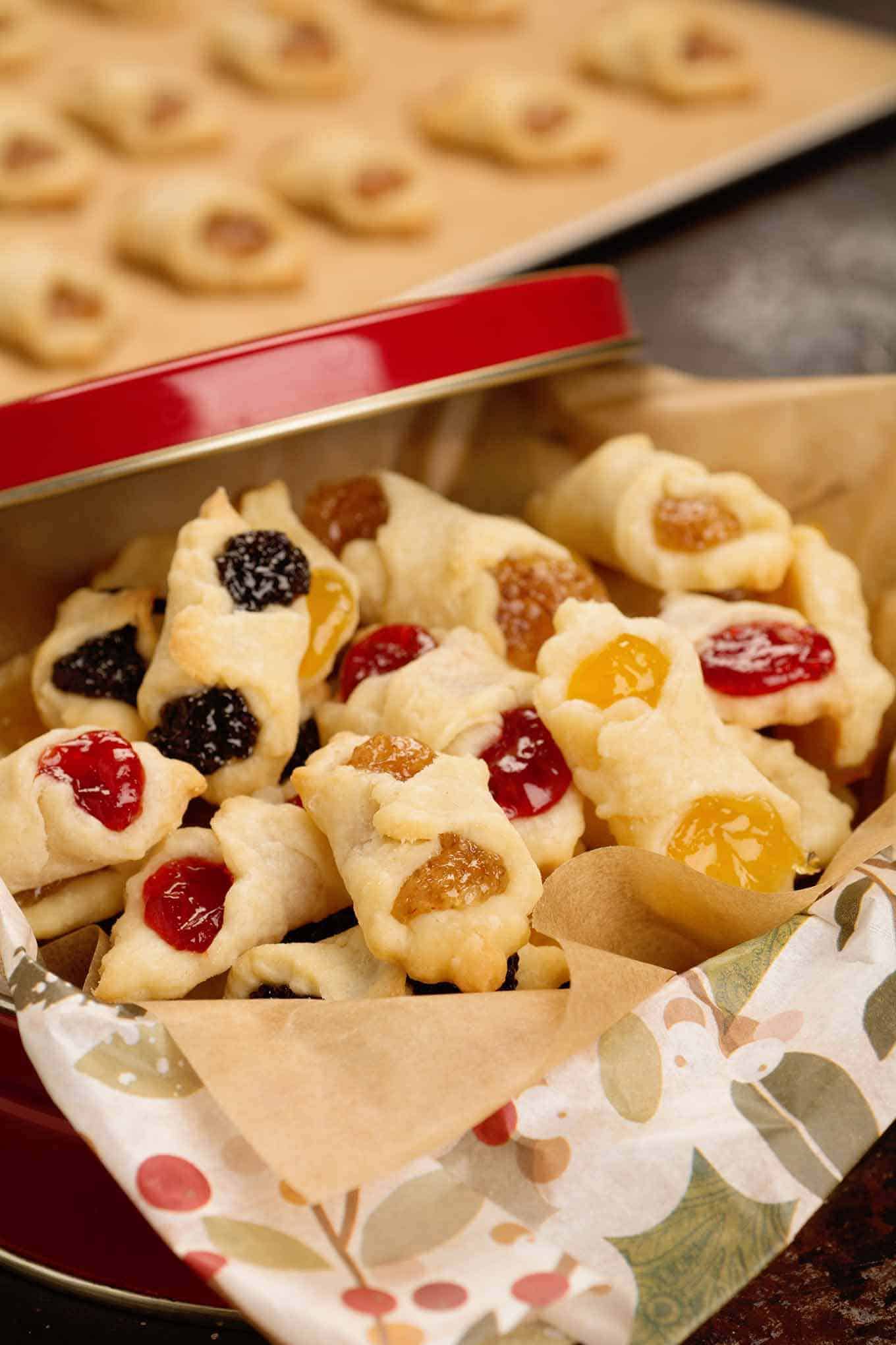 A red Christmas gift tin filled with apricot, almond, poppy seed, and raspberry Hungarian kiffles, a baking sheet with more freshly baked kiffles in the background.