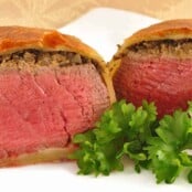 A dinner plate with an individual beef Wellington, sliced in half to show the perfectly cooked beef, topping of mushroom duxelles, and flaky pastry wrap.