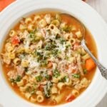 A bowl of Italian chicken soup with ditalini, carrots, escarole, and chicken, sprinkled with grated parmesan cheese and chopped fresh parsley.