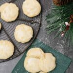 An overhead view of three lemon ricotta cookies glazed and sprinkled with powdered sugar on a linen napkin, a cooling rack with more cookies to the left.