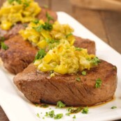A serving platter of pan-seared petite sirloin steaks topped with onion, garlic and pickled banana peppers sautéed in butter - garnished with parsley, tongs in the background.