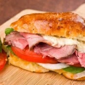A sandwich filled with sliced lamb, tomato, onion, spinach, and roasted garlic thyme mayonnaise on a wooden cutting board.
