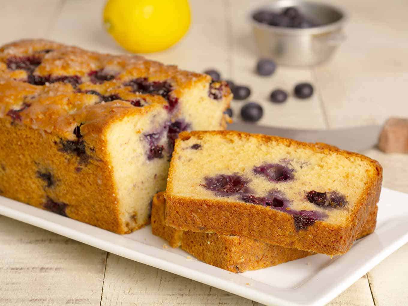 svg%3E svg%3E Slices of lemon blueberry cake on a serving plate with new ingredients within the background. Lemon Blueberry Loaf Cake svg 3E