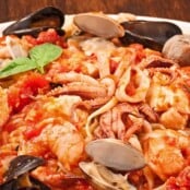 A plate of linguine pasta topped with a medley of seafood including shrimp, mussels, clams, and squid in a rich tomato sauce, garnished with fresh basil leaves.
