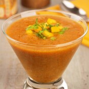A serving of mango gazpacho soup, garnished with chopped mango and cilantro, served in a clear glass with a napkin, spoon, pepper mill, and jar of seasoning in the background.