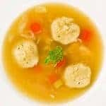 Overhead view of a bowl of matzo ball soup with carrots, celery, shredded chicken, and parsley in clear broth.