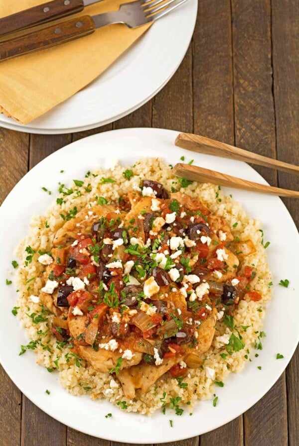 A dinner plate with boneless chicken breasts, topped with a light sauce of tomatoes, Kalamata olives and feta cheese served over a bed of riced cauliflower, extra plates and napkins in the background.