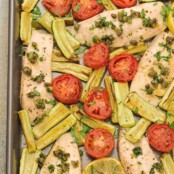 An overhead view of a Mediterranean-style fish dinner on a sheet pan, including tilapia fillets, sliced plum tomatoes, zucchini wedges, capers, and fresh parsley.