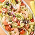 An overhead view of Mediterranean pasta salad with shrimp, artichoke hearts, Kalamata olives, cherry tomatoes, and feta cheese, extra lemon in the background.