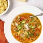 A bowl of hearty minestrone soup with assorted vegetables and pasta topped with grated cheese, and fresh parsley, ditalini and a wedge of Parmesan on the side.