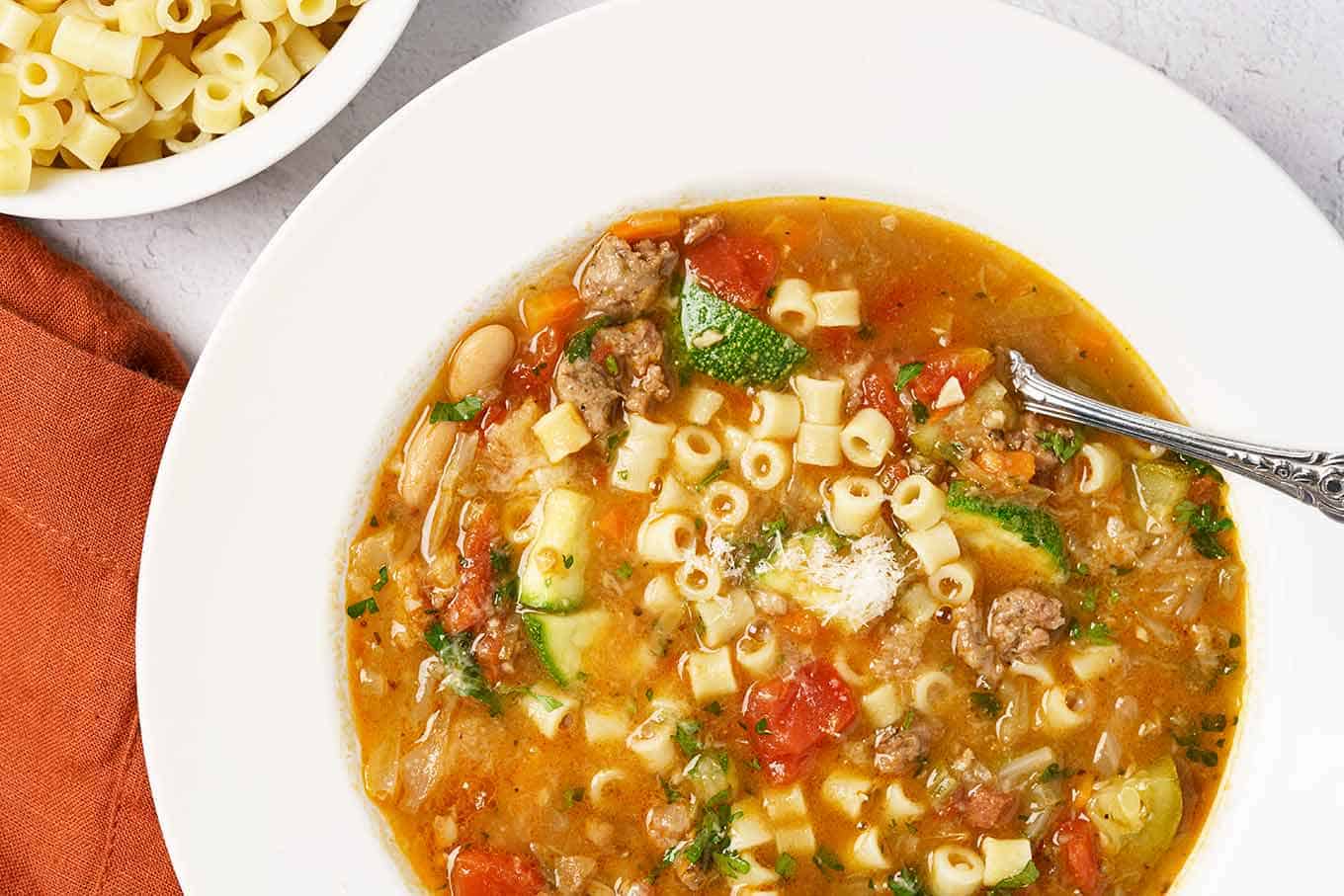 A close view of homemade minestrone soup with visible chunks of Italian sausage, tomatoes, zucchini, beans, cabbage, carrots, pasta, and more.