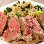Sliced mirin-marinated strip steak topped with sesame seeds, served alongside scallion and kale ramen noodles on a white plate.
