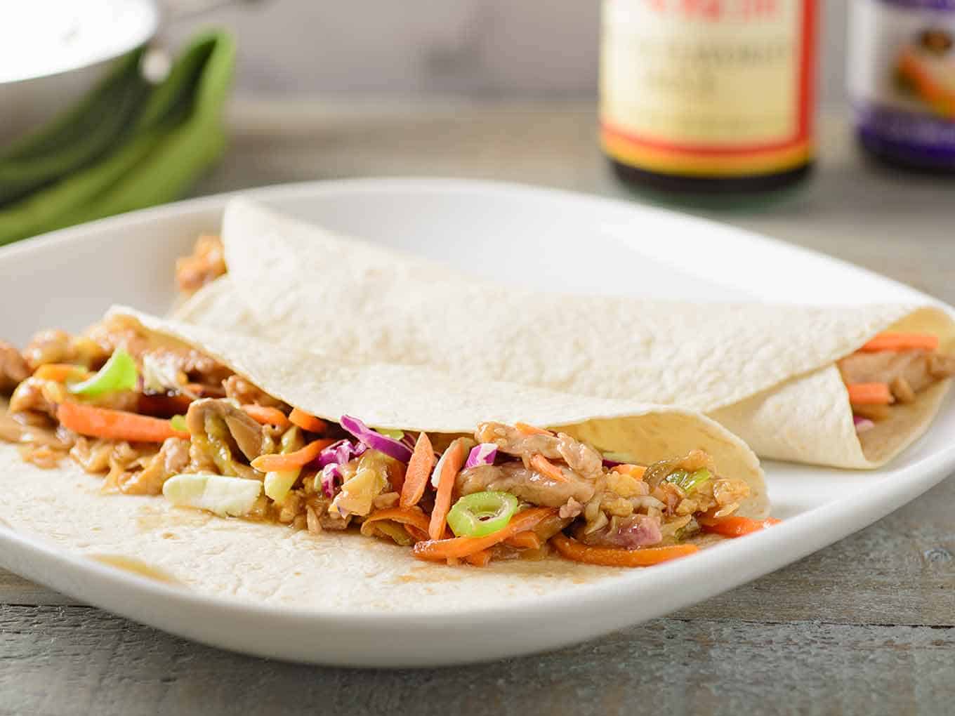 A plate with two thin flour tortillas filled with stir fried moo shu pork, bottles of hoisin and oyster sauce in the background.