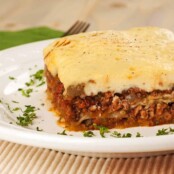 A slice of traditional Greek moussaka made with ground lamb on a white plate, garnished with chopped parsley.