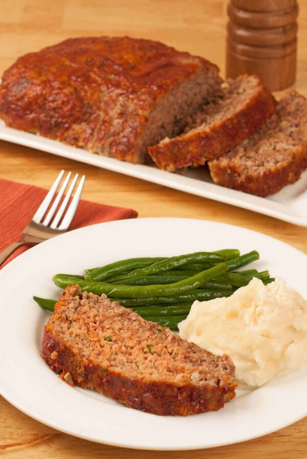 A serving of meatloaf on a dinner plate with mashed potatoes and green beans, the rest of the meatloaf on a serving plate in the background.