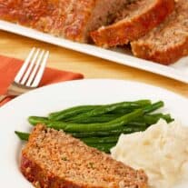 A slice of meatloaf on a dinner plate with mashed potatoes and green beans, a serving platter of meatloaf in the background.