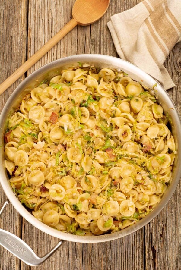 A sauté pan filled with orecchiette pasta tossed with sautéed shredded Brussels sprouts, bacon, onion, and garlic, a wooden spoon and kitchen towel in the background.