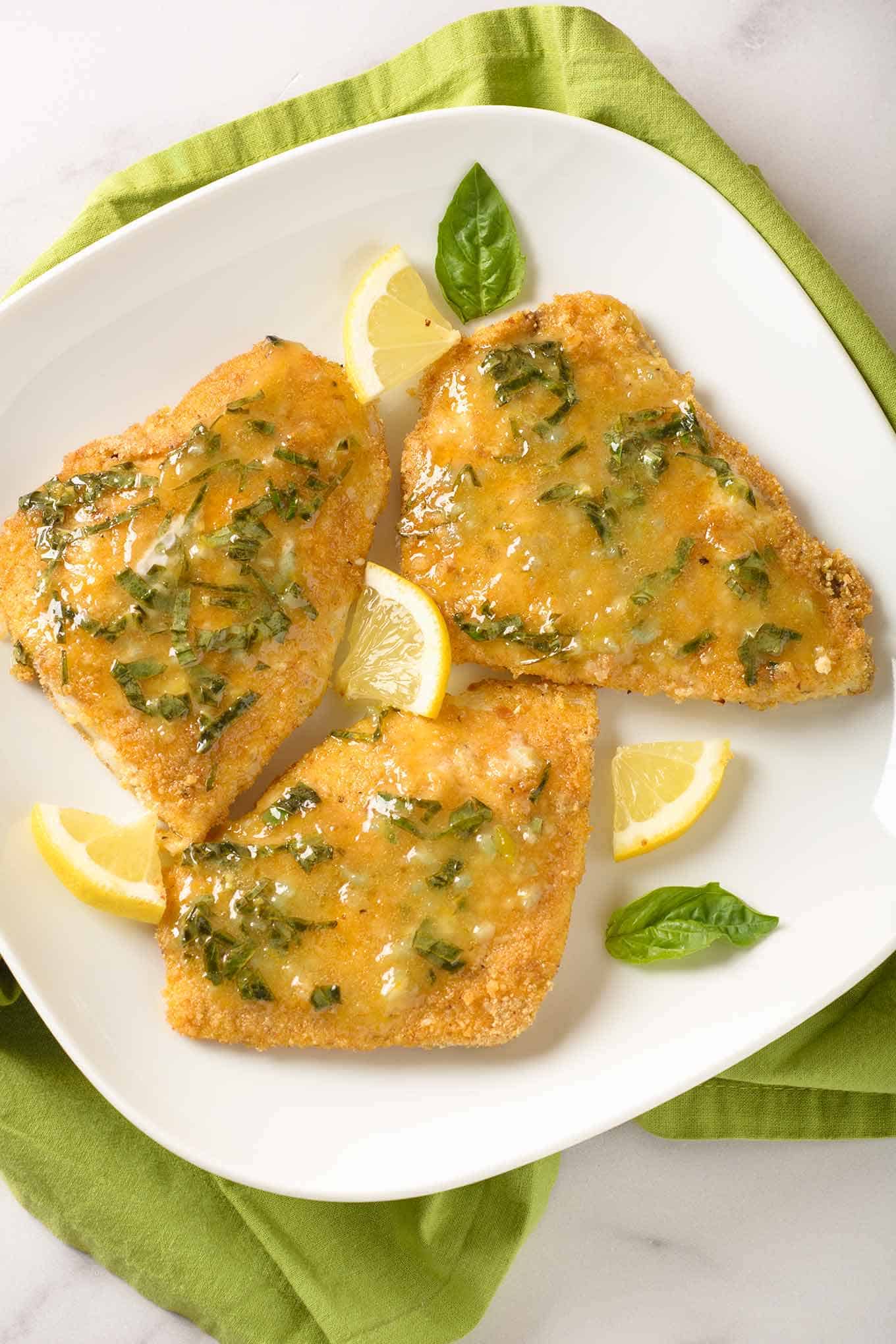Three pieces of breaded, oven-fried fish fillets topped with lemon butter infused with fresh basil and garnished with basil leaves and lemon wedges.