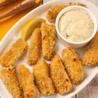 A serving platter of oven-fried fish sticks with a golden crispy coating, served with tartar sauce and lemon wedge.