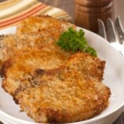 An oval plate with three breaded, oven-fried pork chops, garnished with parsley, a pepper mill and serving fork in the background.