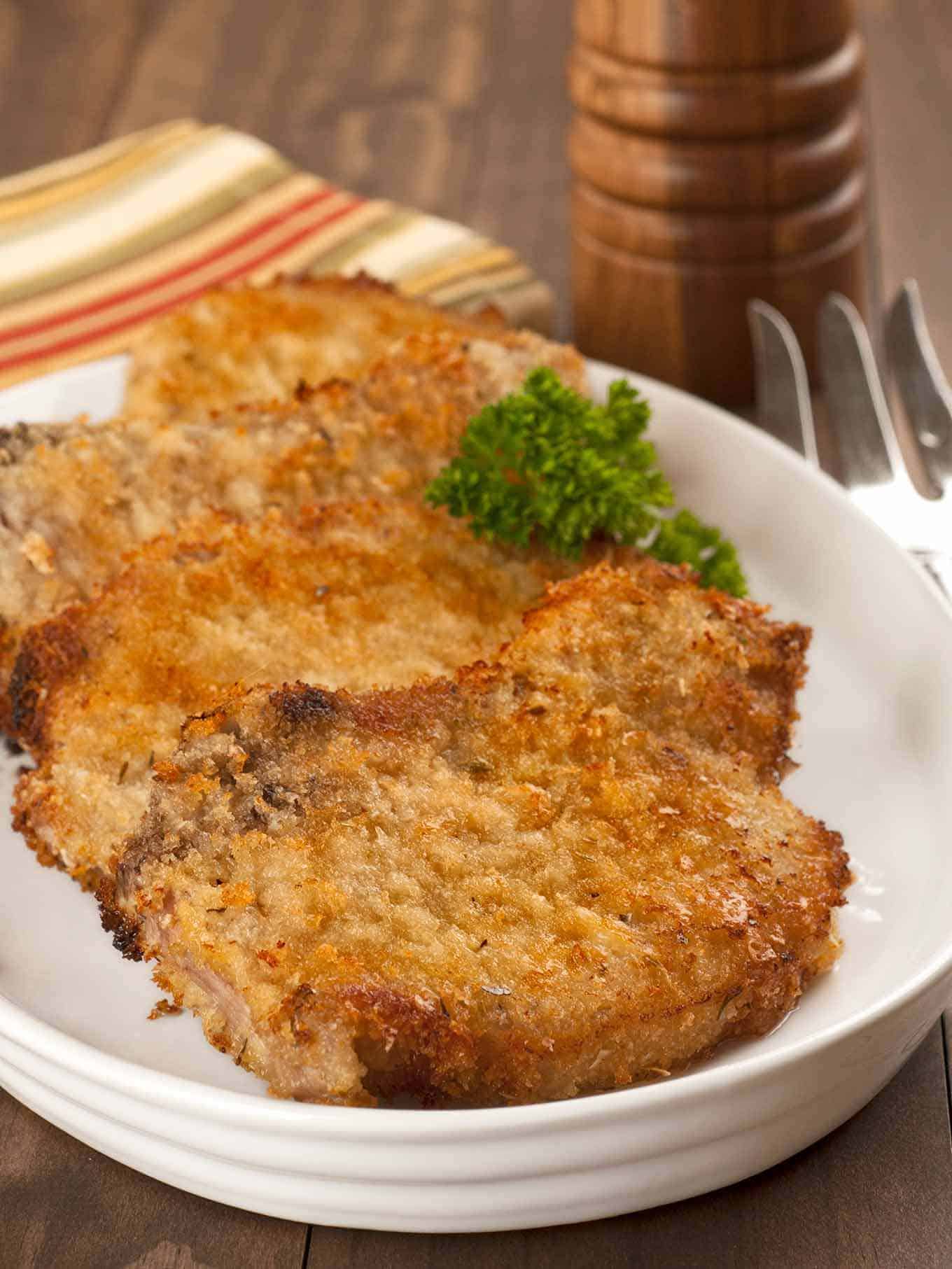 An oval plate with three breaded, oven-fried pork chops, garnished with parsley, a pepper mill and serving fork in the background.