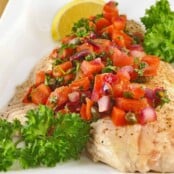 A white plate with oven-roasted snapper fillet topped with a vibrant tomato and caper salsa, garnished with fresh parsley and a lemon wedge.