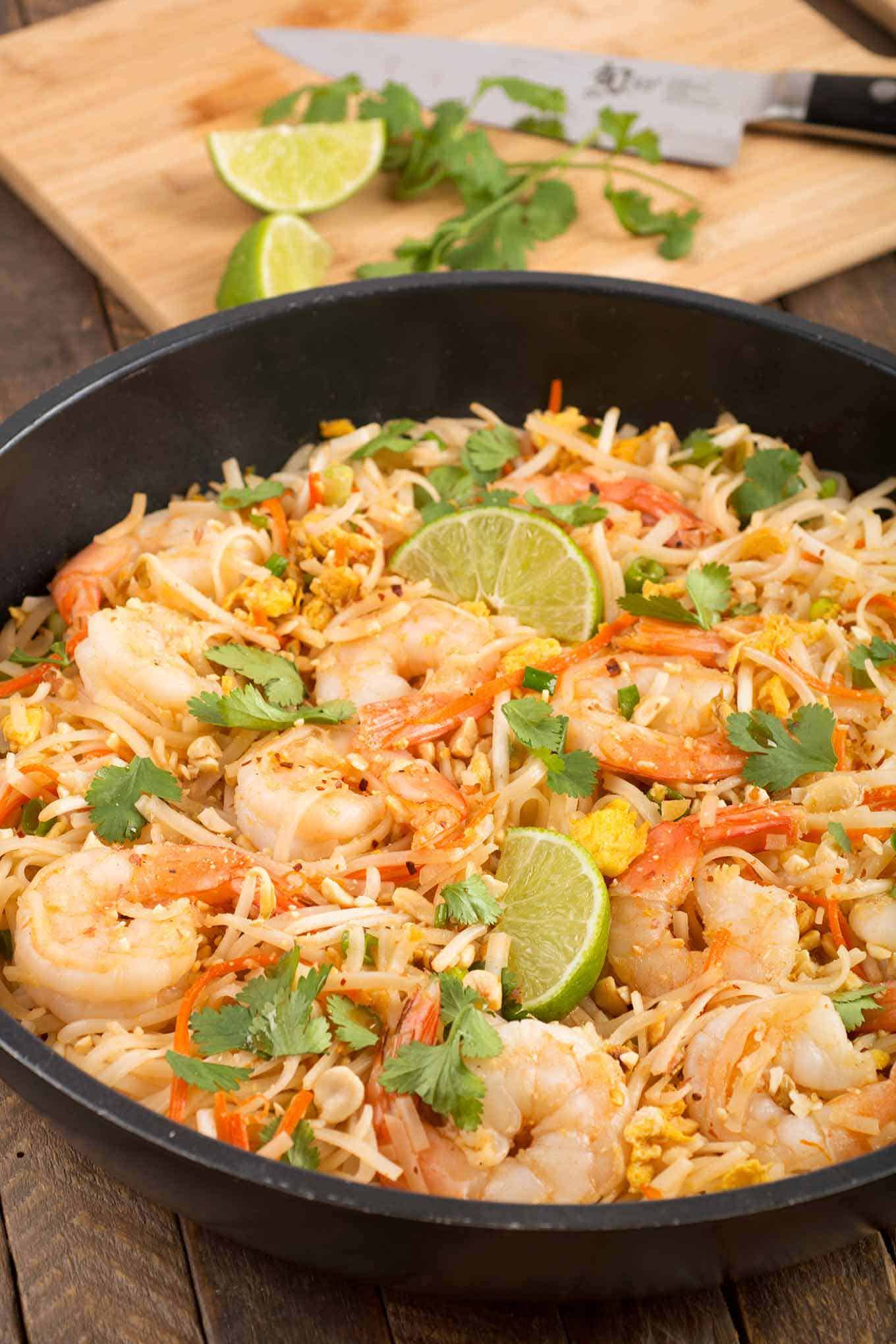 Shrimp Pad Thai in a frying pan garnished with lime wedges and cilantro, cutting board in the background.
