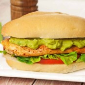 A pan-seared chicken sandwich with avocado mayo, lettuce, and tomato on a sandwich roll, a pepper mill in the background.