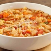 A close-up of a white bowl filled with pasta e fagioli soup, showing pasta, beans, and vegetables in a broth.