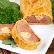 Cheese-stuffed pork tenderloin wrapped in golden puff pastry, served on a white plate garnished with fresh herbs.