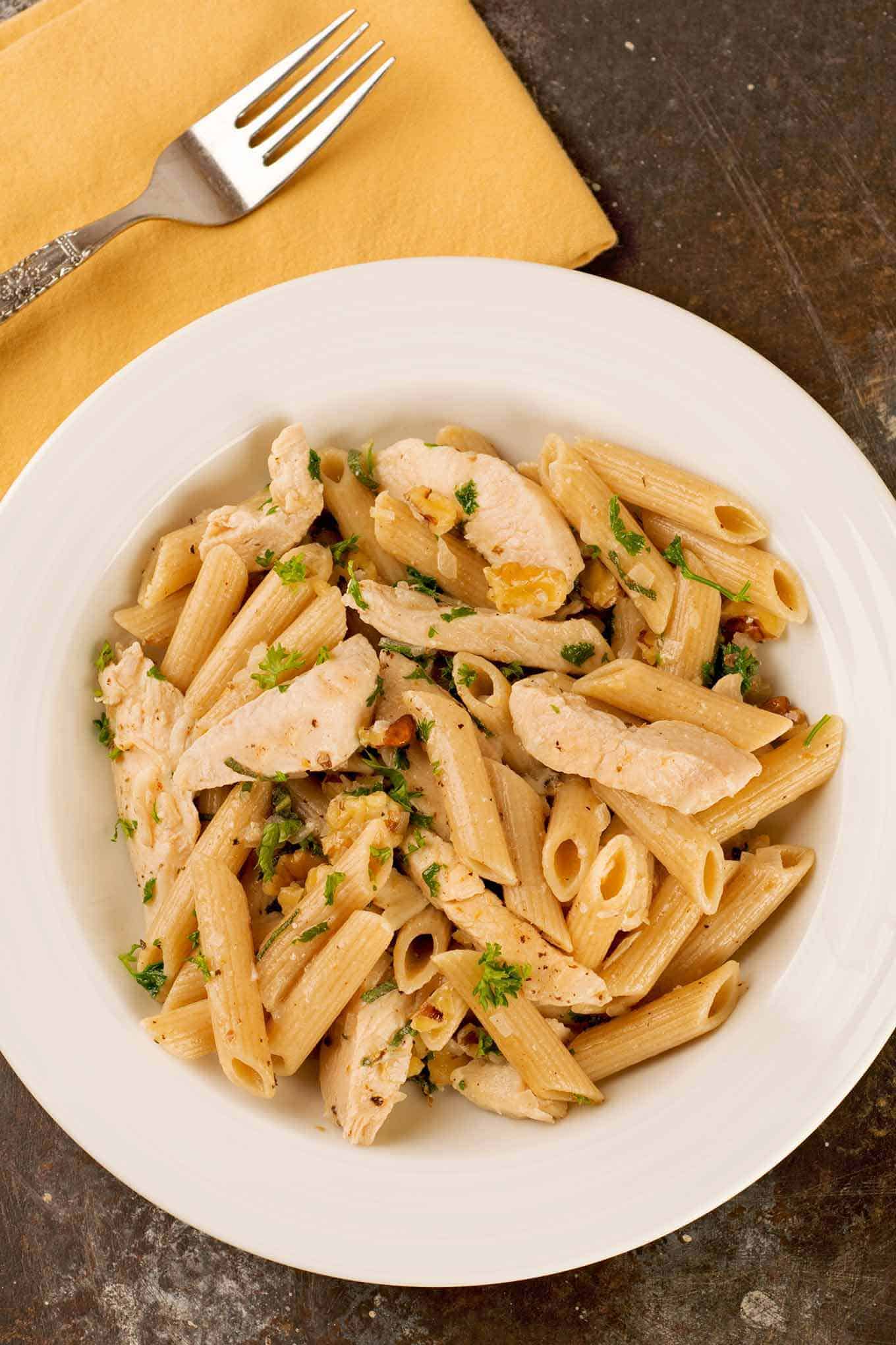 A single serving of penne pasta in sage brown butter sauce with chicken, walnuts, shallots, and a garnish of chopped parsley and ground nutmeg.