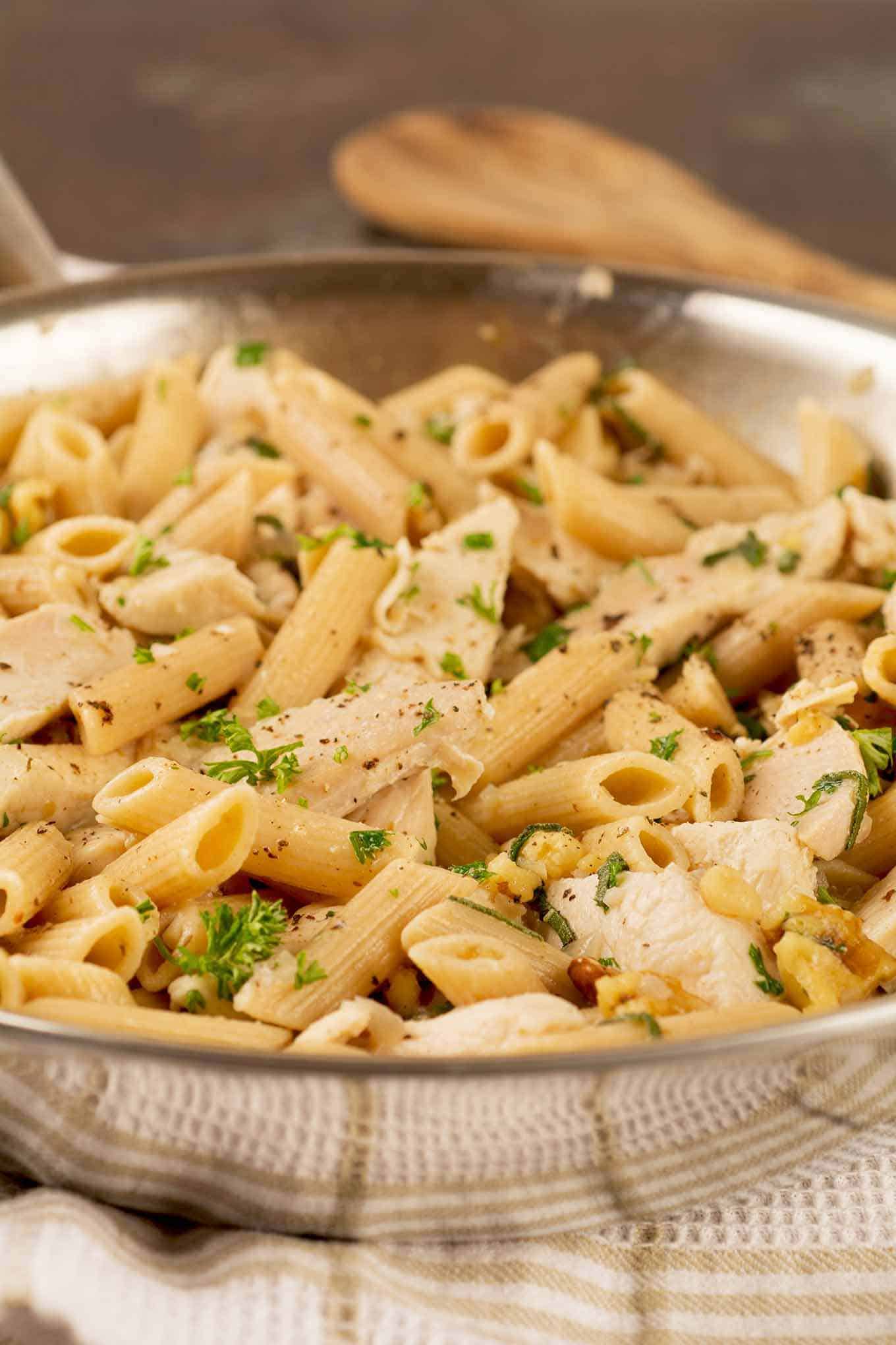 A frying pan filled with penne pasta, chicken breast strips, and toasted walnuts tossed with with brown butter sauce and garnished with parsley.