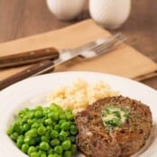 A peppercorn crusted filet mignon topped with compound butter on a plate with mashed potatoes and peas, napkin with fork and knife in the background.