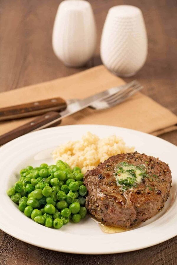 A peppercorn crusted filet mignon topped with compound butter on a plate with mashed potatoes and peas, napkin with fork and knife in the background.