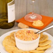 A white crock filled with pimento cheese on a plate surrounded by crackers, a wine bottle, wine glass, and cracker topped with cheese in the background.