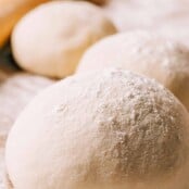 Round balls of pizza dough on a cutting board, dusted with flour, a rolling pin in the background.